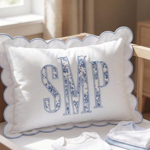 May include: A rectangular white pillow with a scalloped edge trimmed in light blue. The center features the letters SMP in a large, bold font filled with a blue and white floral pattern. Below the pillow are folded white and blue baby bodysuits.