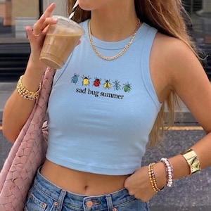 May include: A light blue sleeveless crop top featuring a graphic of six colorful insects arranged in a row above the black text "sad bug summer". The shirt has a ribbed crew neckline and is paired with blue denim jeans and a pink woven shoulder bag.