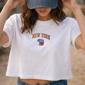 May include: A white short-sleeved crop top featuring an embroidered design on the chest with the text NEW YORK in orange letters above a blue basketball hoop graphic containing an orange basketball. A dark blue baseball cap is also visible.