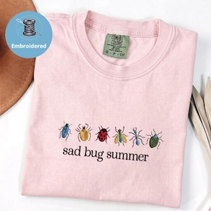 May include: A pale pink t-shirt featuring embroidered insects in multiple colours. The words "sad bug summer" are embroidered beneath the insects. The shirt has a crew neck and short sleeves. The label reads "Comfort Colors".