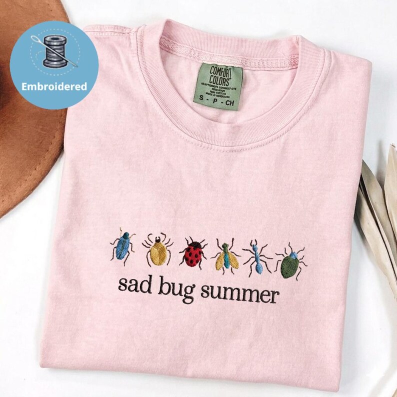 May include: A pale pink t-shirt featuring embroidered insects in multiple colours. The words "sad bug summer" are embroidered beneath the insects. The shirt has a crew neck and short sleeves. The label reads "Comfort Colors".