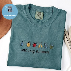 May include: Teal t-shirt with embroidered insect designs and the phrase "sad bug summer." The shirt has a crew neck and a Comfort Colors label. The design includes a variety of colourful bugs.