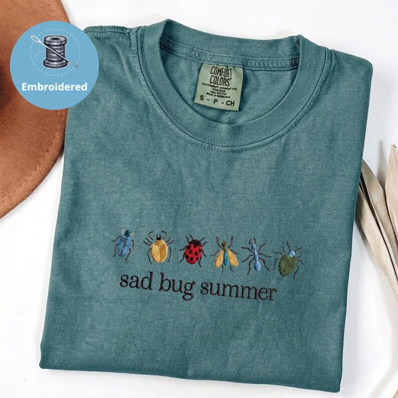 May include: Teal t-shirt with embroidered insect designs and the phrase "sad bug summer." The shirt has a crew neck and a Comfort Colors label. The design includes a variety of colourful bugs.