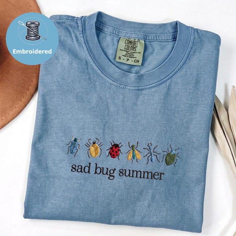 May include: A blue t-shirt with embroidered insects and the phrase "sad bug summer." The shirt has a crew neck and a small tag. The shirt is folded and appears to be made of a soft material.