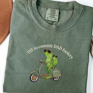 May include: Olive green t-shirt with the text "Off to commit DAD foolery." The shirt features an embroidered design of two green frogs wearing sunglasses riding a green scooter. The shirt has a Comfort Colors tag.