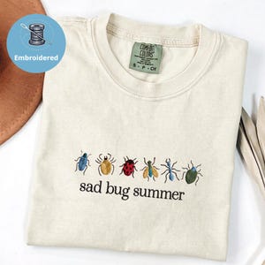 May include: Cream-coloured t-shirt with embroidered insects and the phrase "sad bug summer." The design features a variety of colourful bugs. The shirt has a Comfort Colors tag.