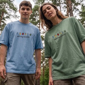 May include: Two people wearing matching t-shirts. One is light blue, the other is green. Both shirts feature an embroidered bug design and the text "sad bug summer" in black.