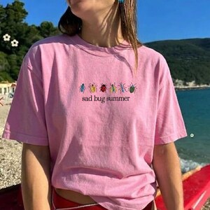 May include: Pale pink short-sleeved t-shirt featuring the phrase "sad bug summer" in black, with six small, colourful insect illustrations above. The shirt is made of a soft fabric, suitable for casual wear.