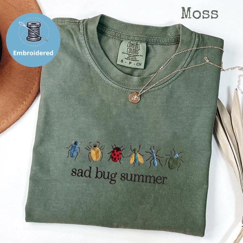 May include: A moss green t-shirt with embroidered insect designs and the text "sad bug summer." The shirt has a crew neck and short sleeves. A silver necklace with a pendant is draped across the shirt.