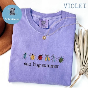 May include: A violet-coloured t-shirt with embroidered insect designs and the text "sad bug summer." The shirt has a crew neck and a small tag. A gold necklace with a pendant is draped across the shirt.