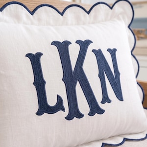 May include: A white rectangular throw pillow featuring a scalloped edge trimmed in navy blue thread. The center of the pillow displays a large, embroidered monogram in navy blue thread with the capital letters LKN in a decorative serif font.