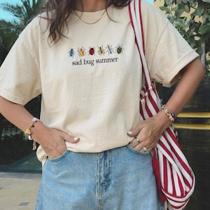 May include: A cream-coloured t-shirt featuring the text "sad bug summer" and bug illustrations in various colours. The shirt has short sleeves and a loose fit. The person is wearing blue denim shorts and carrying a red and white striped tote bag.