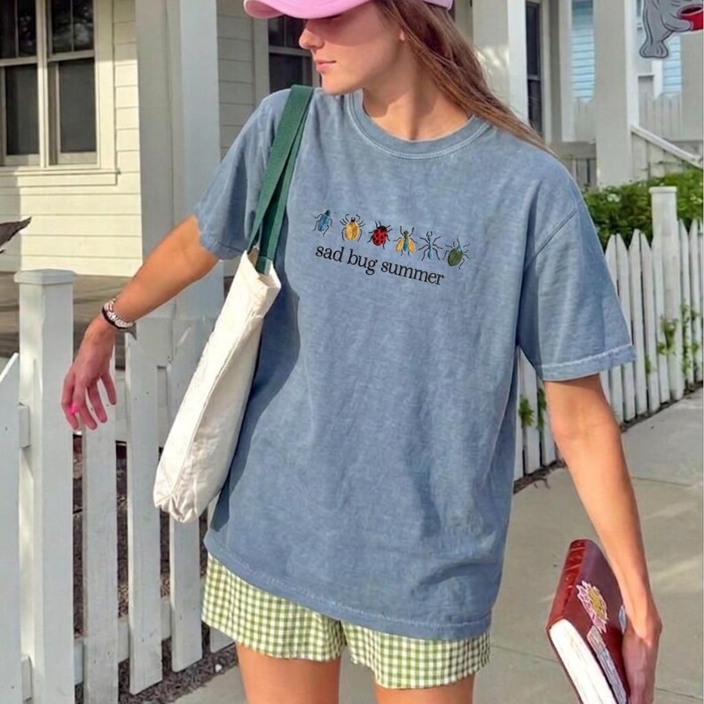 May include: A blue t-shirt featuring the phrase "sad bug summer" and bug embroidery. The shirt is worn with green and white checked shorts. A white tote bag with a green strap is also visible.