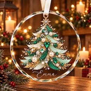 May include: A circular crystal ornament featuring a detailed Christmas tree illustration adorned with green and clear crystals, gold accents, and white pearls. The name Emma is printed in white script at the bottom, and the ornament hangs from a cream ribbon.