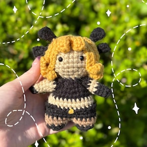 May include: A handmade crochet doll with a bee-like design. The doll features a tan face and body, a black and tan striped skirt, and a black top. The doll has yellow hair and black antennae. The doll is held in a hand against a blurred green background.