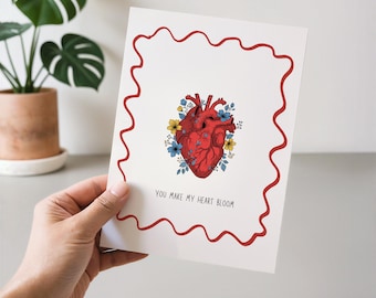 May include: A white greeting card with a red wavy border. The card features a detailed illustration of a red heart surrounded by blue and yellow flowers. The text "YOU MAKE MY HEART BLOOM" is printed at the bottom.