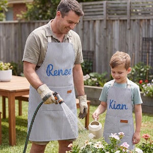 Op de afbeelding: Twee gestreepte schorten, één met de naam "Renee" in blauw geborduurd, de andere met "Kian". Beide schorten hebben een bruin leren label en worden gedragen tijdens het tuinieren.