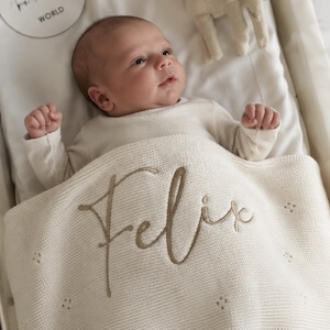 Könnte beinhalten: Eine cremefarbene Strickdecke für Babys mit dem eingestickten Namen Felix in einer großen, braunen Schreibschrift. Die Decke hat ein dezentes Strukturmuster mit kleinen Lochstickereidetails und bedeckt den Unterkörper eines Babys in cremefarbenem Oberteil.