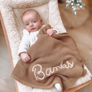 May include: A baby wearing a white long-sleeved shirt is covered by a tan knitted blanket featuring the name Banks embroidered in white cursive script. The blanket has a ribbed border and a textured knit pattern, resting on a cream-colored seat.