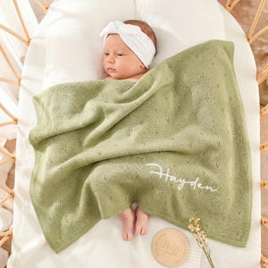 Könnte beinhalten: Ein Baby liegt in einer hellgrünen Decke mit einem zarten Lochmuster. Der Name "Hayden" ist in weißer Schrift auf die Decke gestickt. Das Baby trägt ein weißes Stirnband. Eine Holzscheibe mit der Aufschrift "Hello World" ist ebenfalls zu sehen.