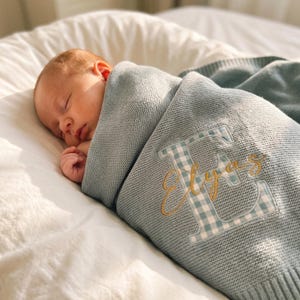 May include: A light blue knitted baby blanket wrapped around a sleeping infant. The blanket features a large, light blue and white gingham letter E embroidered with the name Elyas in gold cursive thread across the centre of the letter.