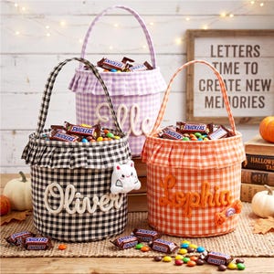 Peut inclure: Trois paniers en tissu à motifs vichy violet, noir et orange, chacun doté d'un bord à volants, d'une anse et d'un nom brodé en écriture cursive. Les paniers sont remplis de barres chocolatées Snickers et de bonbons colorés.