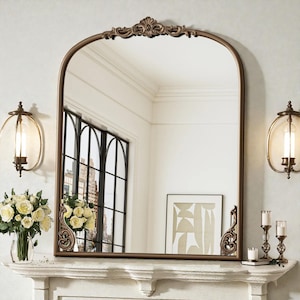May include: An arched bronze-framed mirror with ornate detailing at the top and sides. The mirror reflects a room with a window, artwork, and sconces. A bouquet of white roses and candles are on the mantelpiece below.