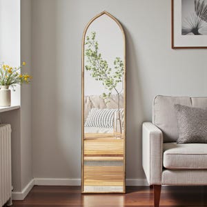 May include: A tall, arched-top floor mirror with a gold-colored frame. The mirror reflects a room with a wooden table, a sofa, and a vase with greenery. The mirror is a decorative home decor item.