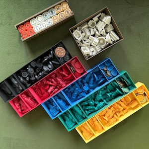 May include: An overhead shot of various board game components organized in colorful trays. The trays are black, red, blue, teal, and yellow, filled with game pieces, tokens, and miniatures. Two additional boxes hold more game pieces.