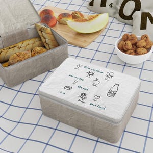 Könnte beinhalten: Eine hellgraue Brotdose mit einem weißen Deckel mit Strichzeichnungen und dem Text "there are things you can buy, the rest is in your heart." Die Box steht auf einer blau-weiß karierten Tischdecke, mit Brot, Obst und Nüssen in der Nähe.