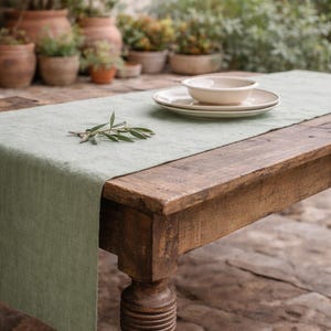 May include: A rustic wooden table with a sage green linen runner. A white bowl and two plates are set on the runner. An olive branch rests on the table. The background features potted plants and a stone patio.