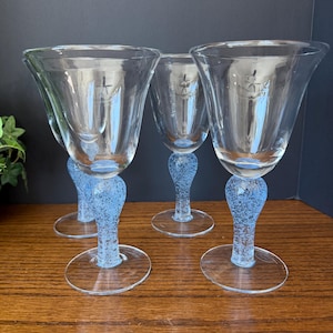 May include: Four clear wine glasses with a light blue, textured stem and base. The bowl of each glass is clear and flared. The glasses are arranged on a wooden surface.
