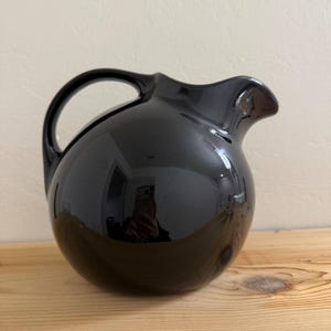 May include: A glossy, spherical black ceramic pitcher featuring a smooth, rounded body, a curved handle on the left side, and a small, integrated pouring spout on the right. The surface is highly reflective, showing a clear mirror-like finish.