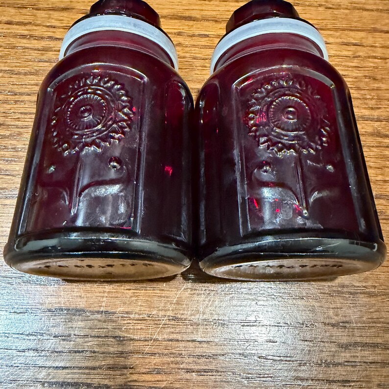 May include: Two vintage, dark red glass jars with white lids. Each jar has a decorative embossed design on the front. The jars are rectangular with rounded shoulders and a slightly tapered shape.