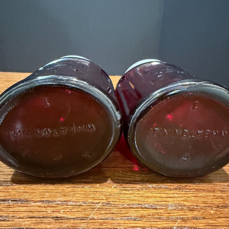 May include: Two antique, dark red glass bottles with rounded bases. The bottoms of the bottles are visible, with the embossed text "WHEATON N.J." in a circular pattern. The bottles are on a wooden surface.