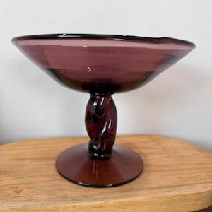 May include: A deep purple glass compote dish featuring a wide, shallow bowl and a thick, twisted stem that connects to a circular base. The glass is translucent with a smooth, polished finish throughout the entire structure of the vessel.