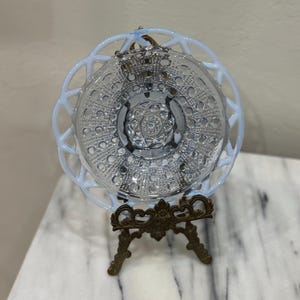May include: A clear glass plate with a delicate, openwork rim and a detailed, textured center. The plate is displayed on a decorative, antique-style brass stand. The plate's rim has a soft, opalescent glow.