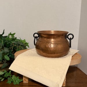 May include: A copper cauldron with two black handles. The cauldron sits on a small wooden stand covered with a cream-colored, patterned cloth. Green foliage is visible in the background. The cauldron has a hammered texture.