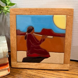 May include: A square tile with a Native American design set in a wooden frame. The tile features a figure in a maroon robe playing a flute, with a blue sky, yellow sun, and desert landscape in the background.