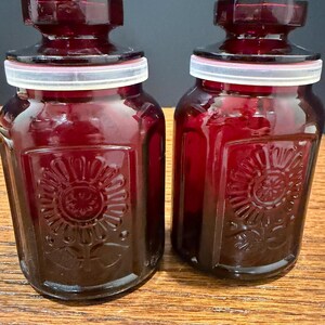 May include: Two ruby red glass jars with lids. Each jar has a square shape with a floral design on the front. The lids are dark red with a white ring. The jars are sitting on a wooden surface.