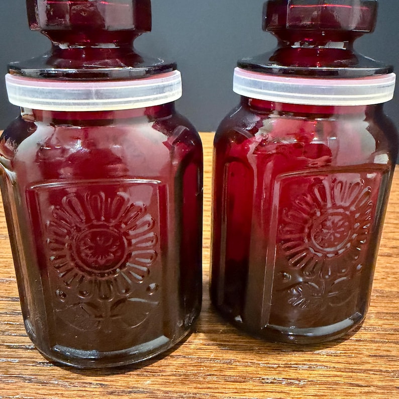 May include: Two ruby red glass jars with lids. Each jar has a square shape with a floral design on the front. The lids are dark red with a white ring. The jars are sitting on a wooden surface.