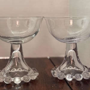 May include: Two clear glass champagne coupe glasses. Each glass has a round bowl, a stem, and a base decorated with small, round glass beads. The glasses are on a dark wooden surface.
