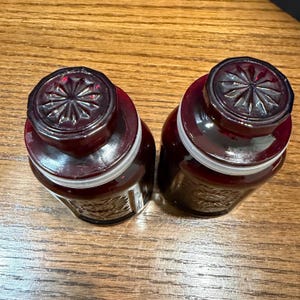 May include: Two matching, dark red glass jars with decorative lids. The jars have a rounded shape and a white band around the middle. The lids are dark red with a starburst design.