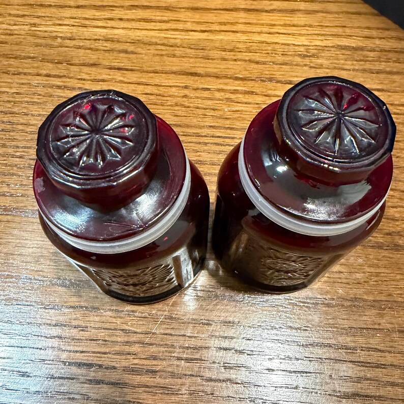 May include: Two matching, dark red glass jars with decorative lids. The jars have a rounded shape and a white band around the middle. The lids are dark red with a starburst design.