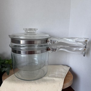 May include: A clear glass double boiler with a lid and a clear handle. The glass pot has a silver metal band around the middle and a clear handle attached with silver metal hardware. The double boiler sits on a wooden surface.
