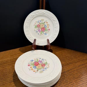 Puede incluir: Una pila de platos de cerámica blanca con un diseño floral en el centro. Los platos tienen un borde en relieve texturizado. Un plato se exhibe en un soporte de madera. El diseño floral incluye flores rosas, amarillas, azules y moradas.