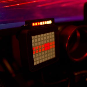 May include: A square digital display unit featuring a grid of illuminated red squares and a horizontal bar of glowing red and yellow lights at the top. The device is encased in a dark frame and emits a vibrant red light against a dark background.