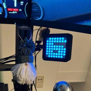 Puede incluir: Una pantalla digital que muestra el número 6 en luz azul. La pantalla está montada en un marco negro. Un pequeño adorno de gnomo con un gorro azul y gris y una barba blanca cuelga cerca. Un monitor que muestra números está en el fondo.