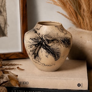 Könnte beinhalten: Eine beige Keramikvase mit einem schwarzen Federdesign. Die Vase steht auf einem Stapel Bücher. Die Bücher tragen die Titel "at 100" und "AUTHENTICS".