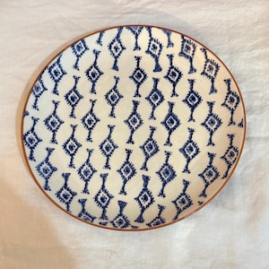 May include: A round ceramic plate with a white background and a repeating blue geometric pattern. The plate has a brown rim. This decorative plate is suitable for serving food.